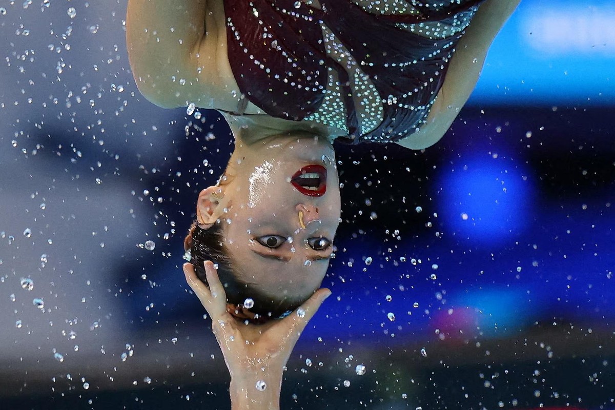 An artistic swimmer performs upside down, with her eyes and mouth open, her head supported by a team member.