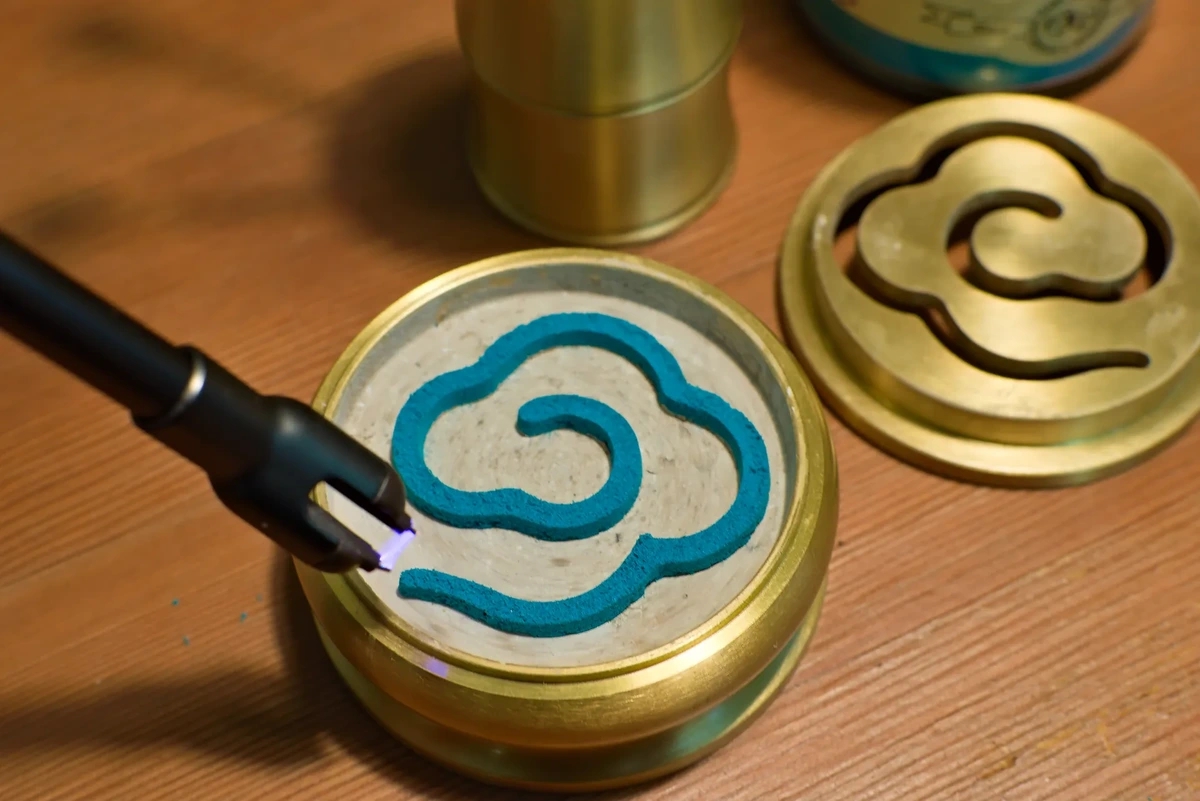 A plasma lighter hovers ominously over the tail of a trail of blue incense powder in the shape of an auspicious cloud, atop a flat layer of ash in a brass censer.