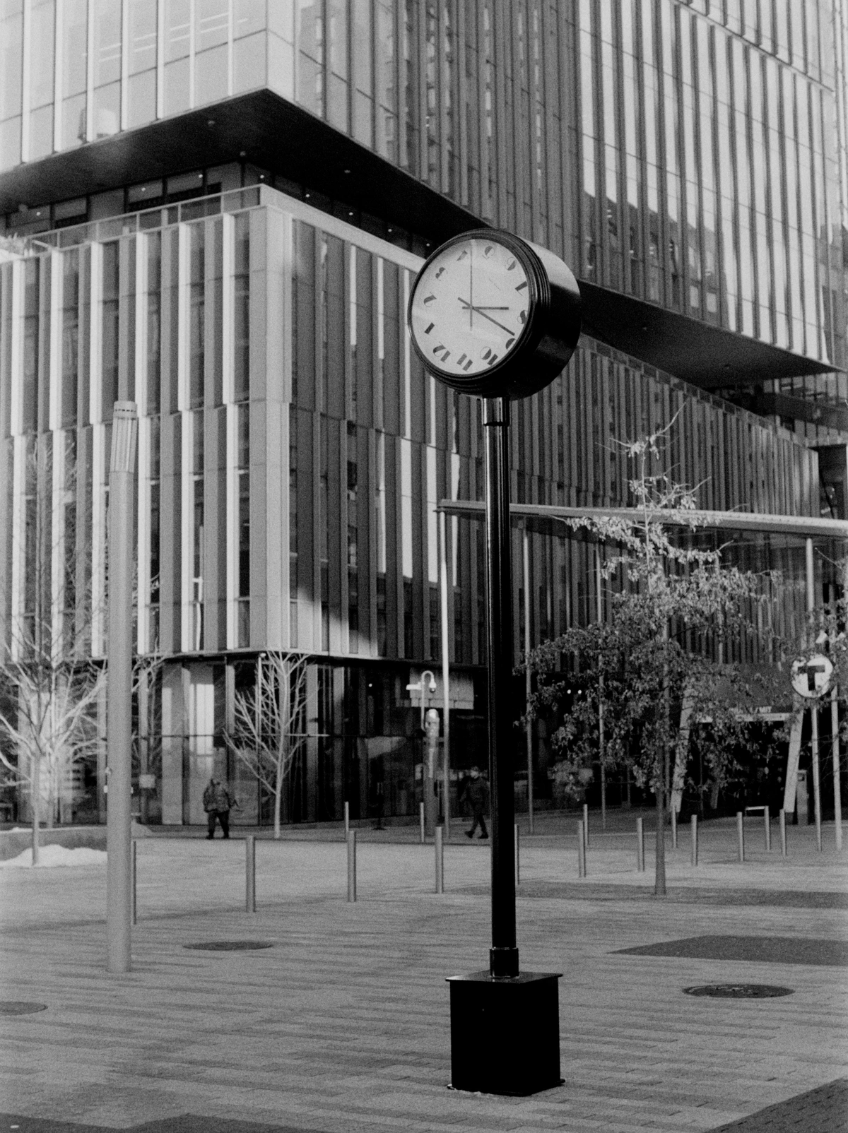 a black and white image showing a plaza pricked with bollards, poles, small trees, and security stations. In the background are some figures walking against the slatted windows of a modern building. In the foreground, an analog street clock stands -- its numbers, confusingly, seem to rotate around the clock hands, each character tilted on its side.   