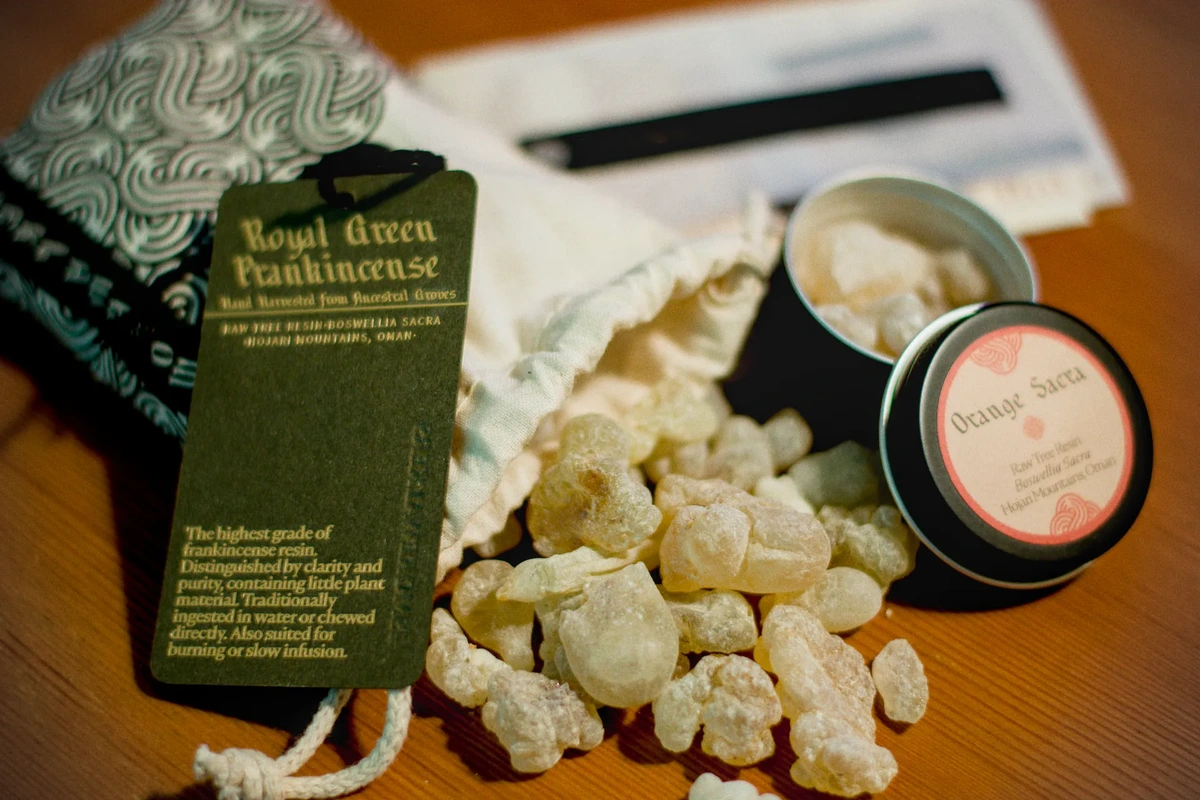 A cloth bag with a drawstring closure with large frankincense tears spilling out of it. A gold-foiled tag with a blind emboss reading "the smokewalkers" describes the frankincense as "Royal Green Frankincense ,hand harvested from ancestral groves, raw tree resin - boswellia sacra, hojari mountains, Oman. The highest grade of frankincense resin. Distinguished by clarity and purity, containing little plant material. Traditionally ingested in water or chewed directly. Also suited for burning or slow infusion." There is also a small tin containing frankincense tears labeled "orange sacra."