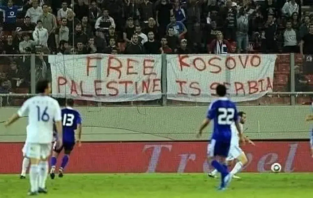 Photo of an ongoing football match with two white sheets over the railings. The first reads 'Free Palestine' and the second 'Kosovo is Serbia', but the way they're positioned makes them read as 'Free Kosovo; Palestine is Serbia'.