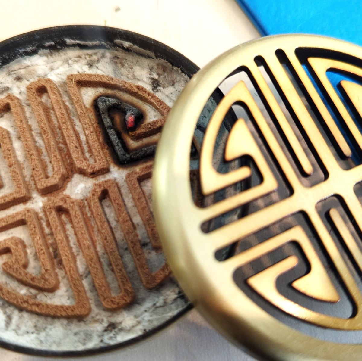 A circular, brass, Chinese-style incense censer with a burning incense seal, incense powder formed via a stencil into a contiguous line of incense that forms an intricate pattern. The seal sits atop white ash specifically made for incense-burning. The censer's brass lid, featuring a cutout resembling an incense seal, is offset atop the censer revealing the seal and ash inside.