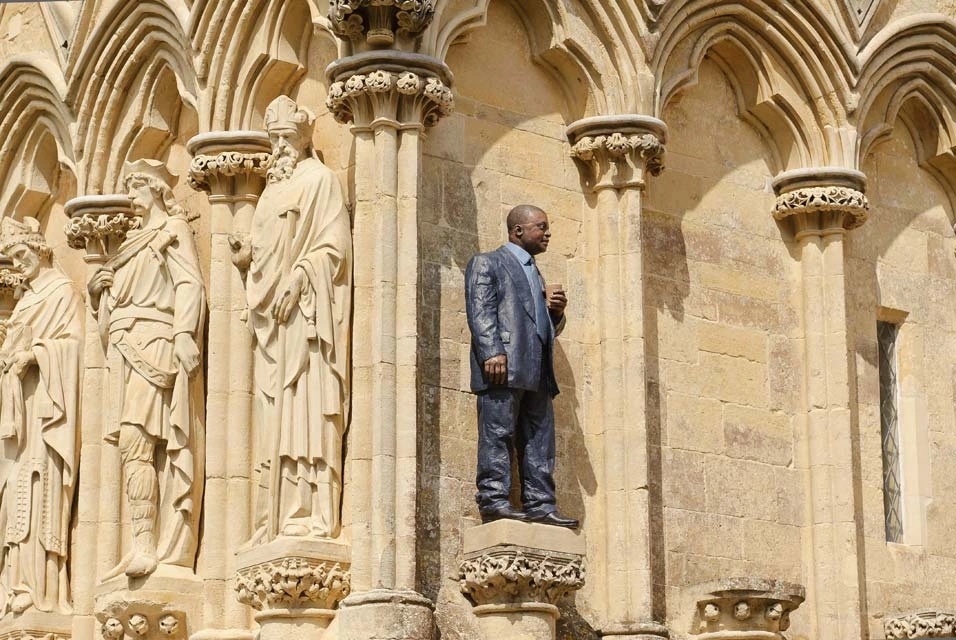 A modern painted sculpture of a man in a suit holding a takeout coffee cup installed alongside early English Gothic sculptures