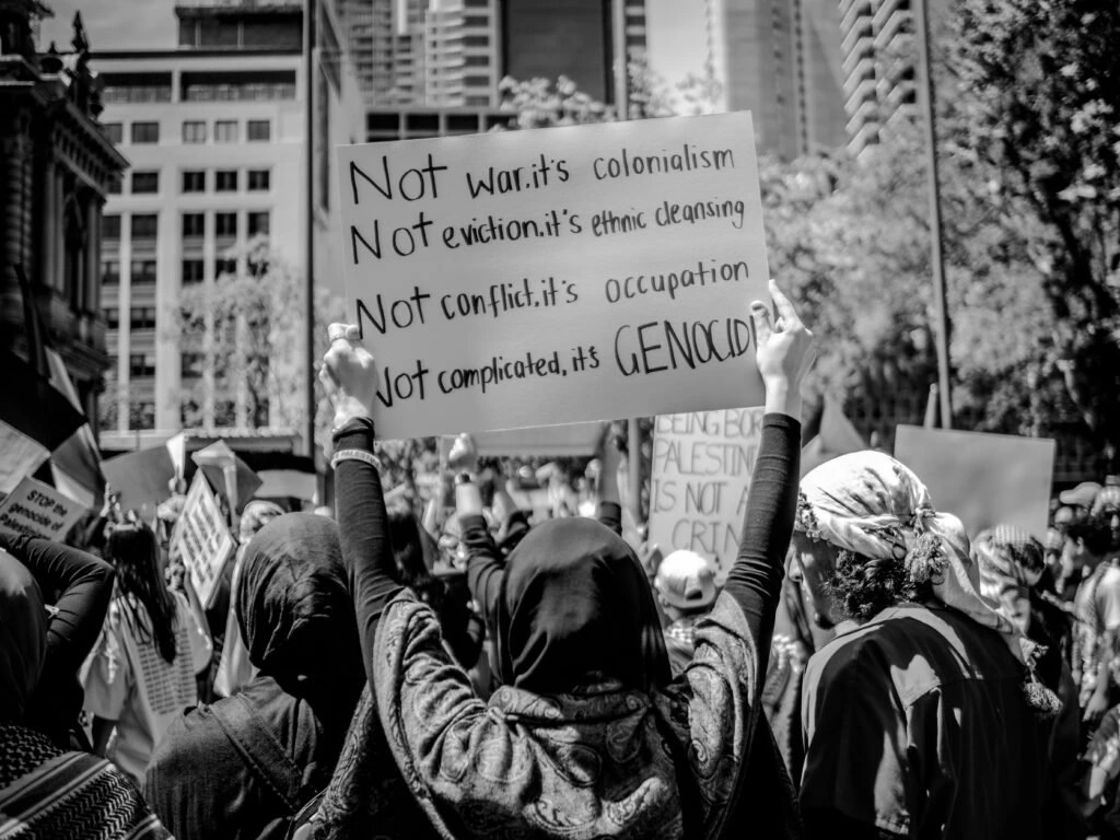 Protesters holding signs at a pro-Palestine rally, with one sign reading “Not war, it’s colonialism. Not eviction, it’s ethnic cleansing. Not conflict, it’s occupation. Not complicated, it’s genocide.”