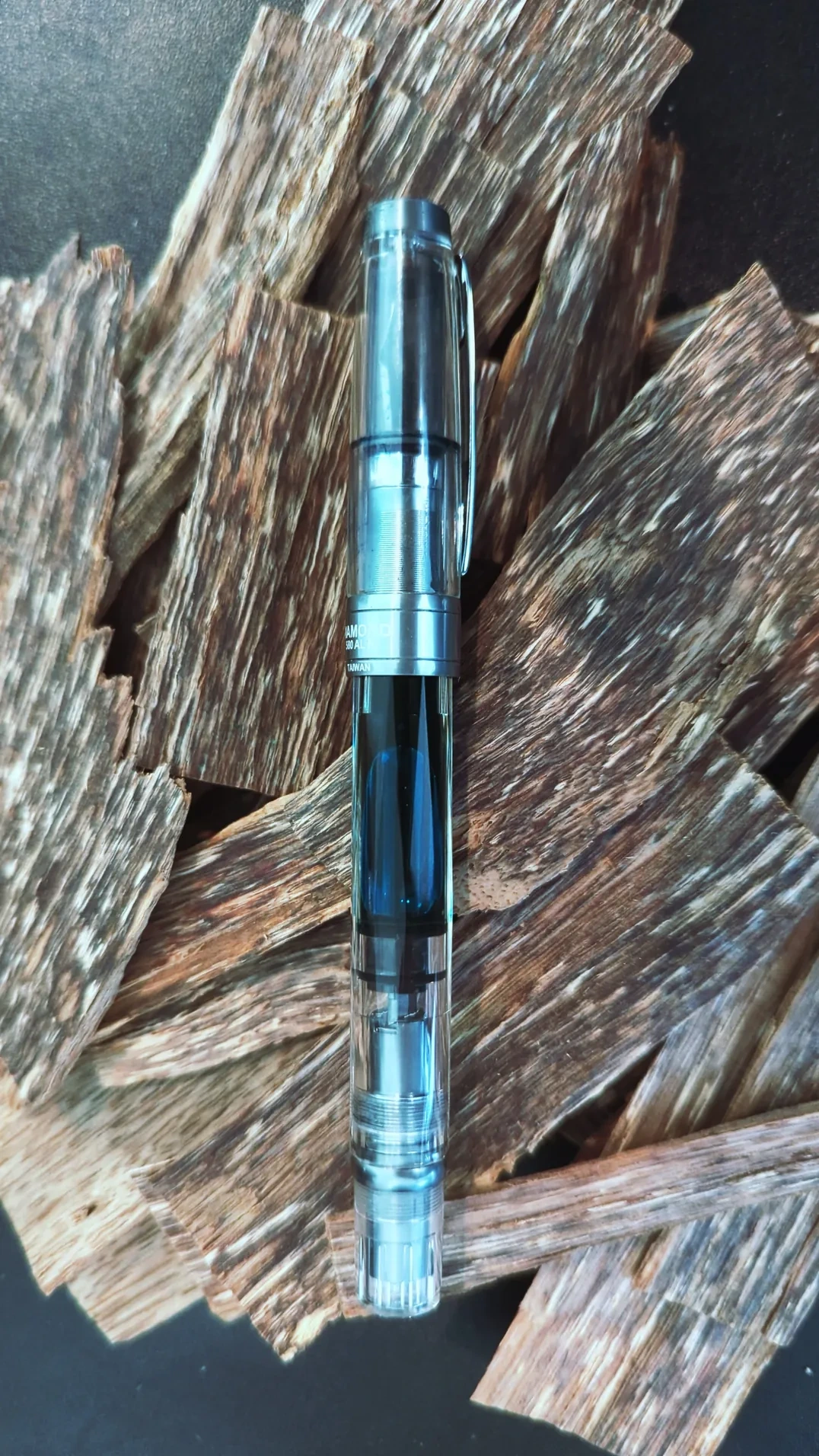 A chunky pen atop a pile of thin stips of agarwood, which are streaked with dark resin. Some pieces are almost as long as the pen, and up to four times as wide.