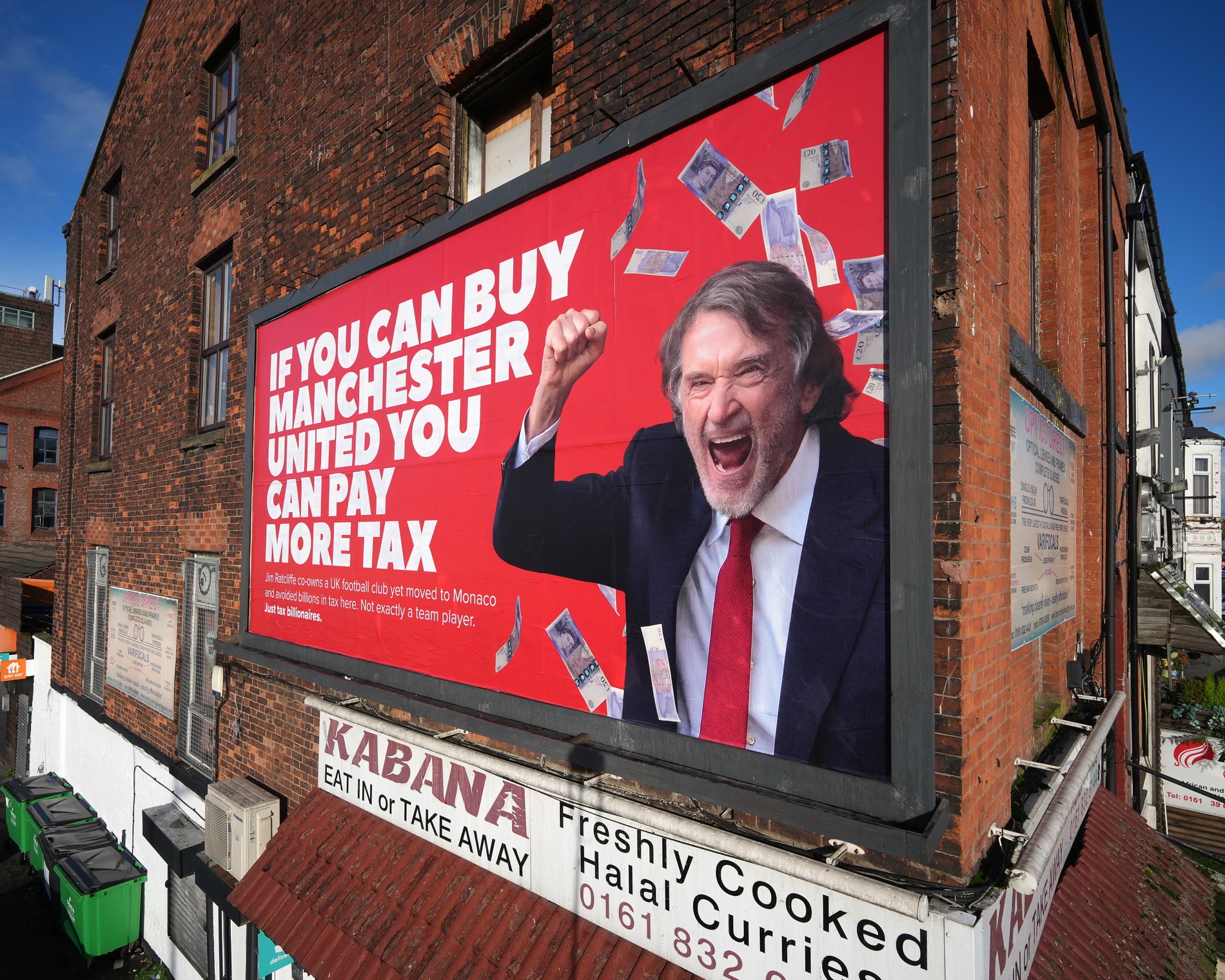 A photo of a billboard on what looks like a building in the UK. The billboard has a red background. On the right of the billboard is a photo of Jim Ratcliffe, a British billionaire, cheering, with £20 notes falling on him. On the left of the billboard, there is text in capital letters saying "IF YOU CAN BUY MANCHESTER UNITED YOU CAN PAY MORE TAX". Underneath that is smaller text, saying: "Jim Ratcliffe co-owns a UK football club yet moved to Monaco and avoided billions in tax here. Not exactly a team player". Underneath that is bold text saying: "Just tax billionaires".