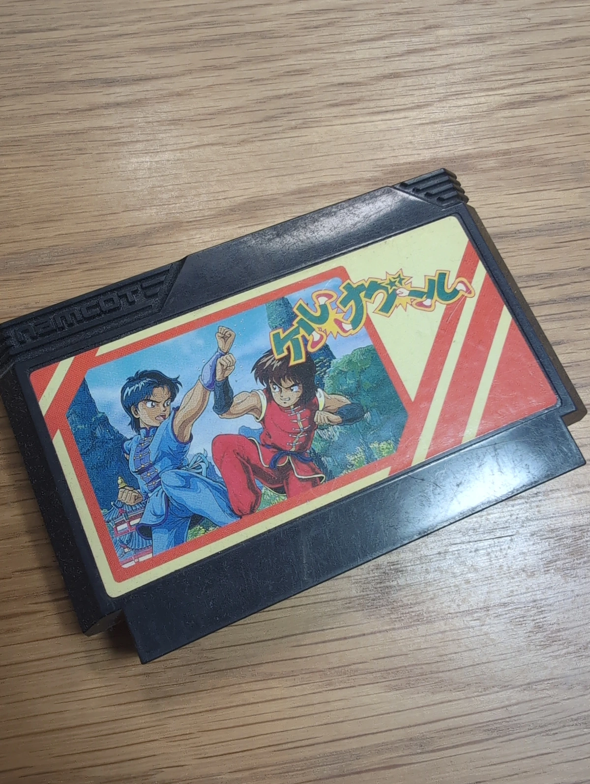 A Famicom cartridge of kerunagūru. It's a black cart with the publisher name "Namcot" physically engraved into it. The art on the cart depicts two asian martial artists in anime style; one dressed in blue and the other dressed in red martial arts gear, fighting in what looks like a forest. 